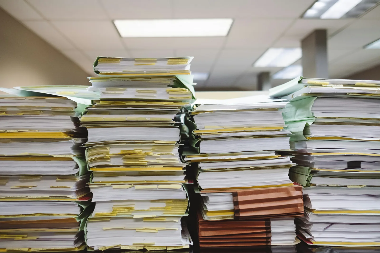 Torenhoge stapels papieren dossiers op een bureau, symbool voor de ongestructureerde papierstroom achter een handmatig inkoopproces.